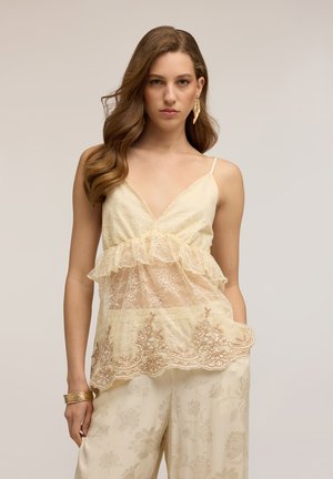Femme aux cheveux bruns ondulés portant un haut en dentelle crème sans manches et un pantalon assorti à motif floral, avec des boucles d'oreilles dorées et un bracelet.