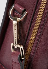 Burgunderrote Lederhandtasche mit einem goldfarbenen Reißverschluss und Beschlägen, die das strukturierte Material und ein Clipverschlussdetail zeigt.