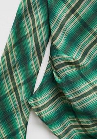 Tessuto a quadri verde e beige con righe orizzontali e verticali incrociate, mostrato drappeggiato e piegato su uno sfondo bianco.