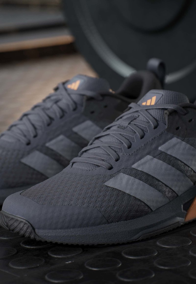 Para ciemnoszarych butów sportowych Adidas z czarnymi paskami i sznurówkami, umieszczonych na teksturowanej czarnej powierzchni.