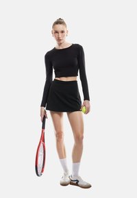 Sort langærmet cropped top parret med en sort nederdel. Holder en grøn tennisbold og et rødt tennisbat. Hvide ankelstrømper og sneakers.