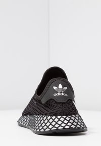 Czarne sportowe buty z siateczki o teksturowanym, przypominającym sieć wzorze, z srebrnym akcentem na pięcie i miękkim, elastycznym kołnierzem.