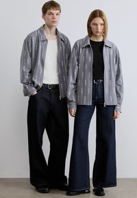 Γκρι πουκαμίσου τζάκετ με κατακόρυφο λευκό κείμενο, συνδυασμένα με μαύρα και σκούρα μπλε παντελόνια φαρδιάς γραμμής. Λεπτό ύφασμα και άνετη εφαρμογή.