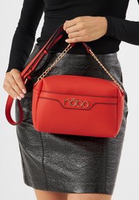 Borsa in finta pelle rossa con texture e ferramenta dorata, caratterizzata da un logo e dettagli con catena. Tracolla regolabile per versatilità.