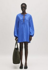 Blaues Langarmkleid mit einem Krawatten-Ausschnitt, strukturiertem Stoff, lockerer Passform und einer Länge über dem Knie, kombiniert mit einer dunkelgrünen Tasche.