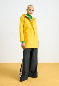 Chaqueta de lluvia impermeable amarilla con capucha, dos bolsillos frontales y cierre de presión. Combinada con pantalones de pierna ancha negros con rayas blancas.