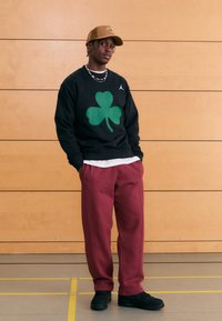 Μαύρο φούτερ με μεγάλο πράσινο τριφύλλι και λογότυπο Jordan, σε συνδυασμό με μπορντό φόρμα και μαύρα αθλητικά παπούτσια, σε ξύλινο φόντο.