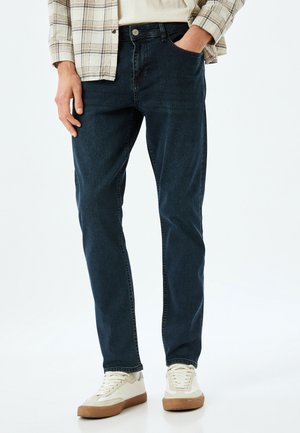 Vaqueros de mezclilla azul oscuro con corte recto, que presentan bolsillos delanteros y cremallera. Combinados con zapatillas de deporte crema y una camisa de cuadros.