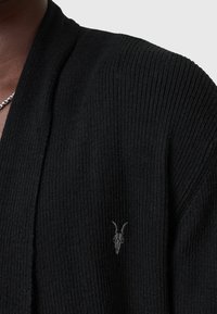 Gros plan d’un pull noir en maille côtelée avec un petit logo brodé de crâne de chèvre sur la poitrine et un collier en chaîne argentée partiellement visible.