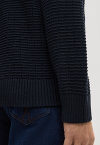 Dunkelblauer, gestrickter Pullover mit einem strukturierten Rippenmuster und gerippten Bündchen, gezeigt in der Nähe einer Hand und Jeans.