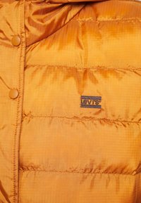 Veste matelassée en tissu satin orange avec un petit patch logo Levi's bleu et noir et des boutons arrondis. Texture lisse et brillante.