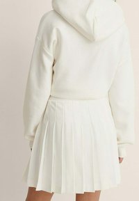 Valkoinen huppari, jossa on huppu ja pitkät hihat, yhdistettynä valkoiseen tyyliin taitettuun hameeseen. Sileä materiaali, minimalistinen muotoilu ja puhtaat linjat.