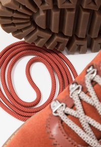 Gros plan sur une botte de randonnée orange avec des lacets beiges, un lacet rouge enroulé, et une semelle de botte robuste et texturée marron sur fond blanc.