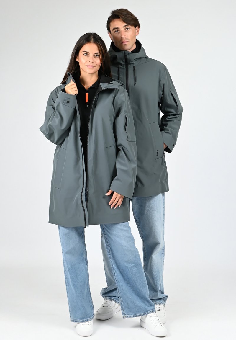 Vestes imperméables grises avec capuches, dotées d'une fermeture éclair à l'avant et de poches latérales, portées sur des hauts noirs, associées à des jeans bleu clair.