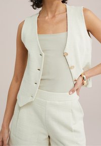 Ljust grönt texturerat väst med guldfärgad knappar, bärs över en ribbad topp, i kombination med matchande byxor. Slät tyg, ärmlös design.