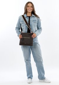 Jonge vrouw draagt een spijkerjasje en spijkerbroek, witte sneakers, glimlacht en houdt een zwarte crossbodytas vast met een voorvak met rits.