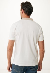 Uomo con capelli corti e scuri che indossa una polo bianca con colletto beige e jeans blu, visto di spalle su sfondo bianco.