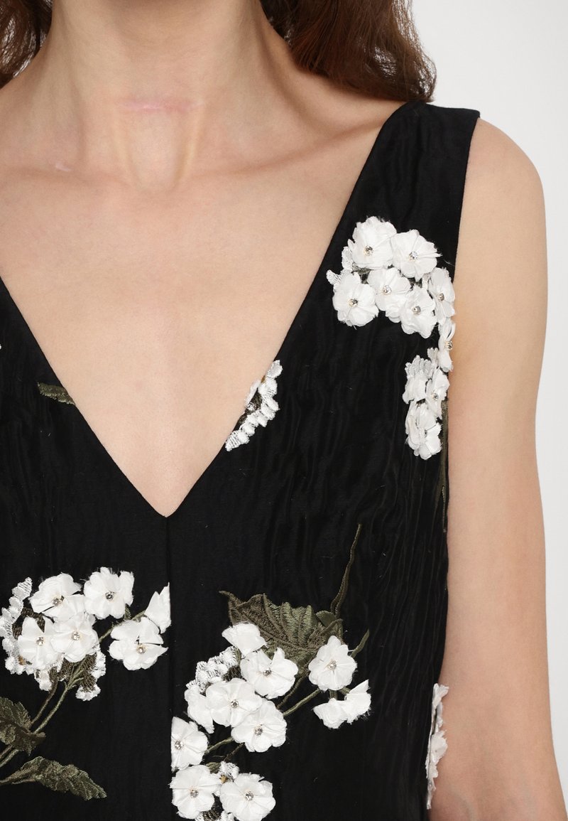 Robe noire avec un décolleté en V profond, ornée de broderies florales blanches et de feuilles vertes. Le tissu texturé rehausse le design.