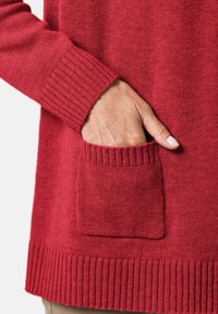 Roter Strickpullover mit geripptem Halsausschnitt und Bündchen, einer vorderen Tasche und einem strukturierten Körper. Der Stoff wirkt weich und warm.