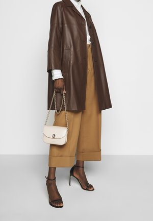 Personne portant un pantalon marron à jambes larges, un chemisier blanc avec des poignets à volants, un manteau en cuir marron foncé, des sandales à talons hauts noires, tenant un petit sac à main beige.