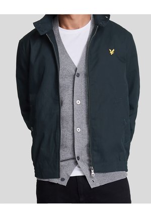 Mand iført en sort lynlåstrøje over en grå knap-cardigan og en hvid T-shirt med hænderne bag ryggen på en ensfarvet baggrund.
