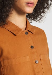 Mujer con cabello oscuro y rizado vestida con una camisa de botón color naranja quemado con dos bolsillos en el pecho, frente a un fondo claro.