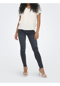 Weißes kurzärmeliges Blusenshirt mit strukturiertem Muster und Kordelzug am Hals; kombiniert mit dunkelblauen Skinny Jeans und Riemchensandalen mit hohen Absätzen.