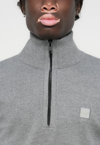Grauer Reißverschluss-Pullover aus Strickmaterial mit hohem Kragen, schwarzem Reißverschluss und quadratischem Logopatch auf der Vorderseite.
