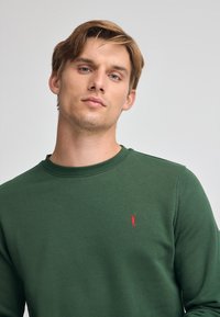 Grön crewneck sweatshirt tillverkad av texturerat tyg, med en liten röd logotypdetalj på bröstet samt ribbade ärmslut och halsringning.