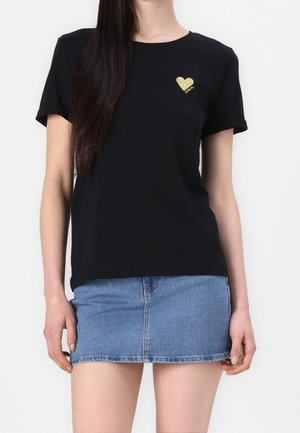 Frau trägt ein schwarzes T-Shirt mit kleinem goldenem Herz und Schriftzug, kombiniert mit einem blauen Jeans-Minirock, vor weißem Hintergrund stehend.