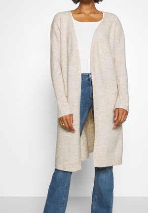 Person trägt eine lange, beige Strickjacke mit offenem Schnitt, ein weißes Stricktop und blaue Jeans und steht vor einem einfarbigen Hintergrund.