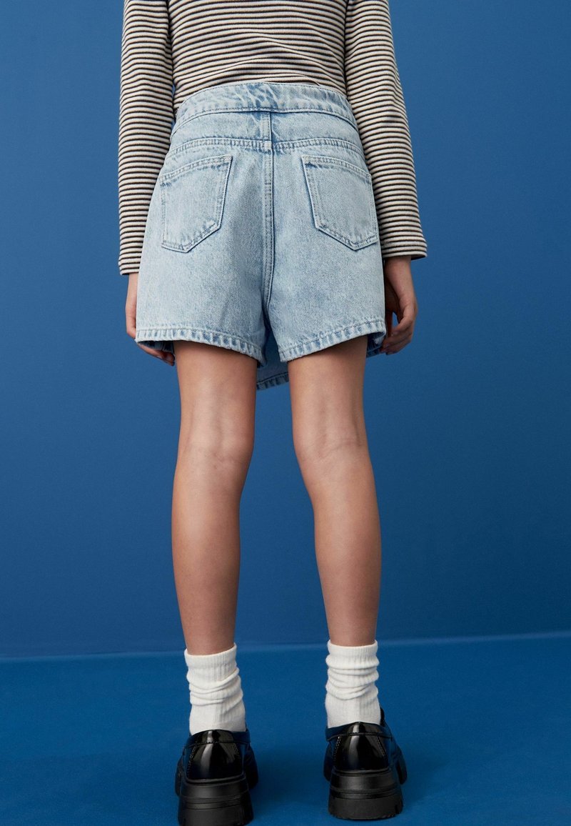 Personne portant un short en jean bleu clair, des chaussures noires avec des chaussettes blanches, et une chemise à manches longues rayée, debout devant un fond bleu.