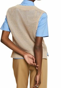 Personne portant un pull sans manches en tricot beige sur une chemise légère à manches courtes bleue et un pantalon couleur camel, avec les mains jointes derrière le dos.