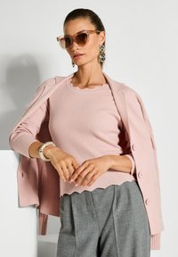 Frau mit Sonnenbrille, großen Kettenohrringen, einem rosa gerippten Oberteil mit gewellten Rändern, grauen plissierten Hosen und einem rosa Mantel über den Schultern drapiert.