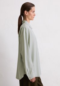 Light green, long-sleeve blouse in a loose fit, featuring a button-down collar and a single chest pocket, against a neutral background.