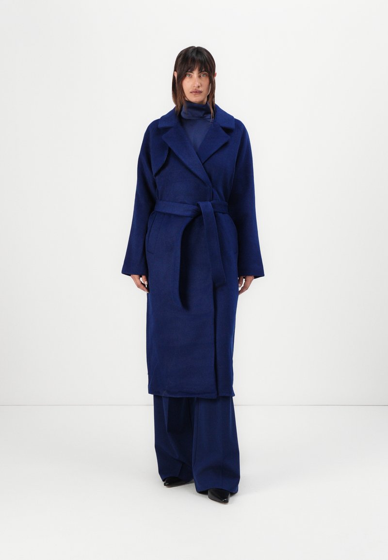 Long blue coat made of textured fabric, featuring a collar, wide sleeves, and a matching belt. Loose-fitting trousers and black shoes.