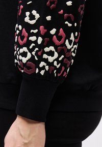 Mustal dressipluusi varrukas soonikkoes mansetiga, millel on valged ja tumeroosad reljeefsed leopardimustrid.