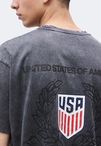 Νεαρό άτομο που φοράει ξεθωριασμένο μαύρο μπλουζάκι με το λογότυπο ασπίδας των ΗΠΑ και το κείμενο "United States of America" στην πλάτη.