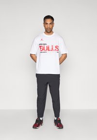 Λευκό κοντομάνικο μπλουζάκι με το γράφημα «Chicago Bulls» σε κόκκινο και μαύρο. Συνδυάζεται με μαύρο παντελόνι με στενή εφαρμογή και παπούτσια με κόκκινες λεπτομέρειες.