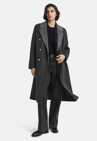 Dubbeldraperad grå ullrock med stor slag, guldknappar och houndstooth-mönster, kombinerad med mörka jeans och svarta skor.