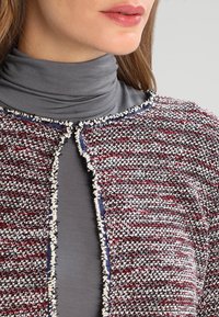 Cardigan tissé avec un motif rouge, blanc et bleu, tissu texturé, devant ouvert et bords bruts. Superposé sur un col roulé gris.