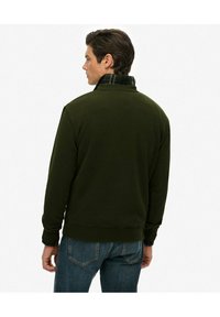 Sudadera de forro polar verde oscuro con un cuello a cuadros, puños acanalados y dobladillo; combinada con jeans azules. Detalles de costura visibles.