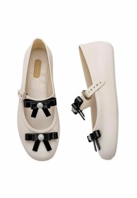 Bailarinas de color beige con lazos negros brillantes y un acento de perla. Tienen una puntera redonda y una correa con una hebilla de metal. Textura suave.