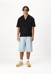 Camisa negra de manga corta con dos grandes bolsillos, pantalones cortos de mezclilla azul claro y zapatillas con detalles amarillos. Unas gafas de sol negras completan el conjunto.