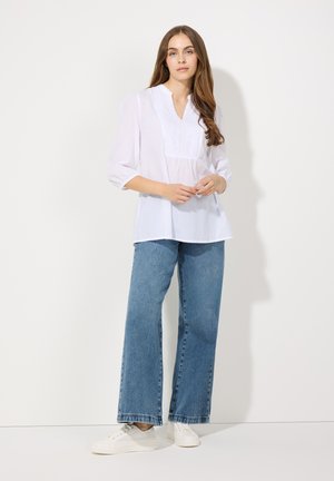 Witte, lange mouwen blouse met een voorflap en geborduurde details, gecombineerd met wijde blauwe spijkerbroeken en witte sneakers.