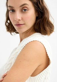 Joven mujer con cabello corto castaño y ojos azul claro, que lleva una blusa de punto blanca sin mangas, mirando suavemente hacia la cámara.