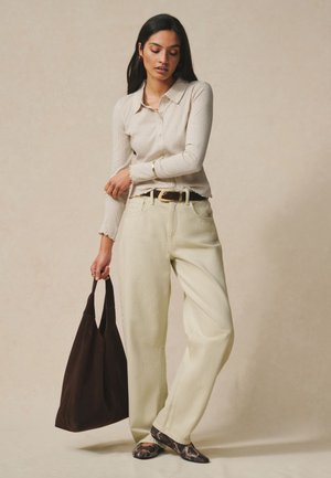 Kvinde i beige ribbet button-up top og brede cremefarvede bukser holder stor mørkebrun taske, iført flade sko med slangeprint.