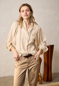 Femme aux cheveux châtain clair portant un chemisier beige à rayures avec des manches bouffantes et un pantalon camel, debout à l'intérieur près de sculptures en bois.