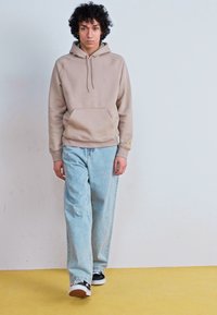 Beige hooded sweatshirt with front pocket, paired with light blue loose-fit jeans and black sneakers. Model stands against a light background.