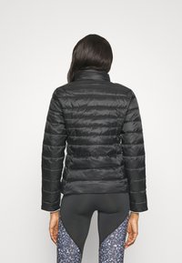 Vrouw gekleed in een zwarte doorgestikte jas en zwarte legging met blauwe luipaardprintpanelen, staand met haar rug naar de camera tegen een effen achtergrond.